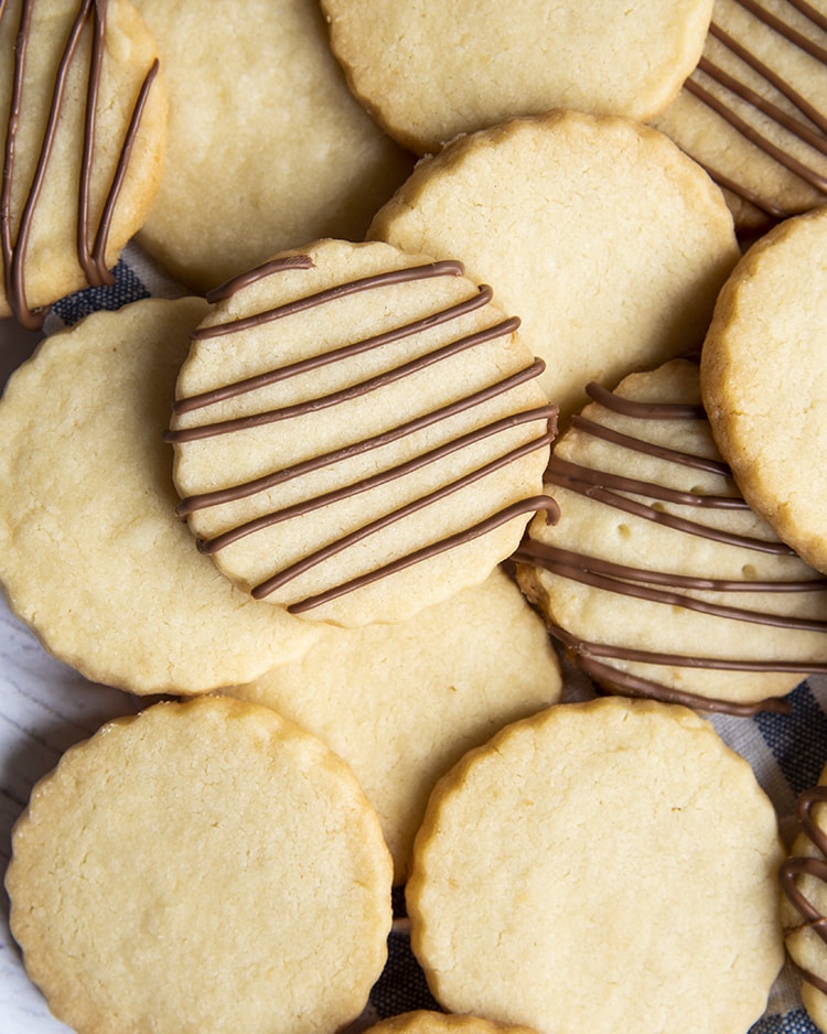 A close up of a pile of shortbread cookies, some of the cookies have chocolate drizzled over the top.
