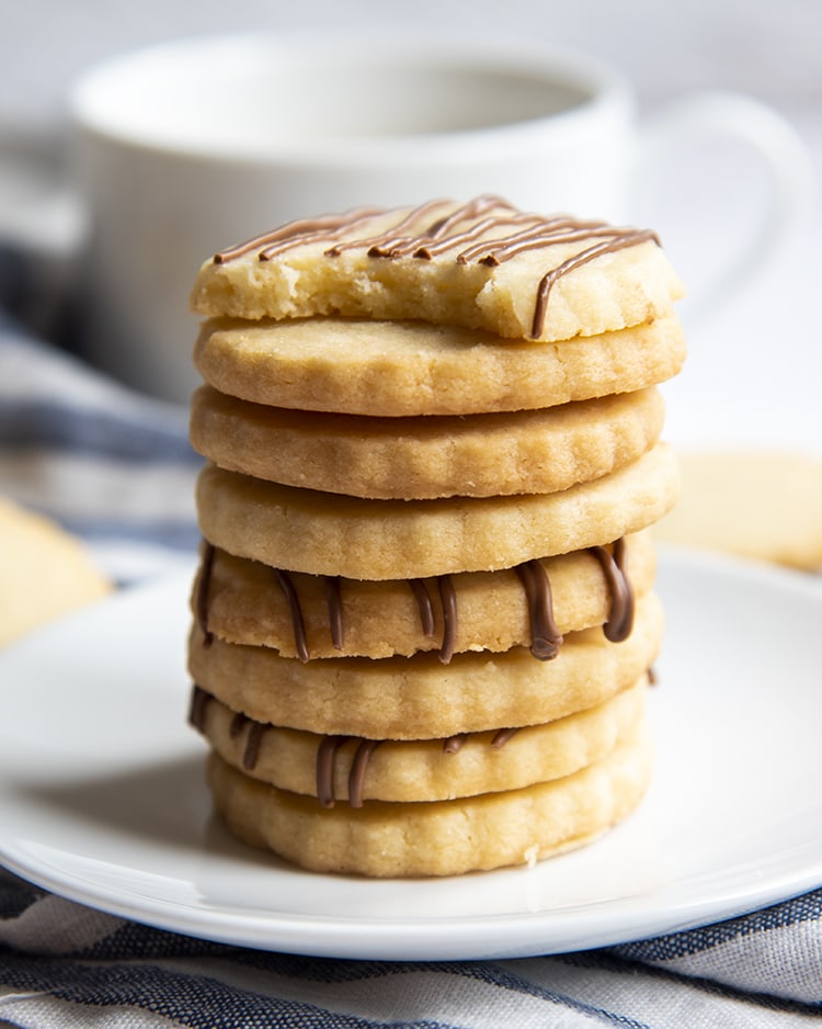 A stack of shortbread cookies with a crimped edge. A few of the cookies have chocolate drizzled over the top. The top cookie has chocolate drizzled, and a bite out of it.