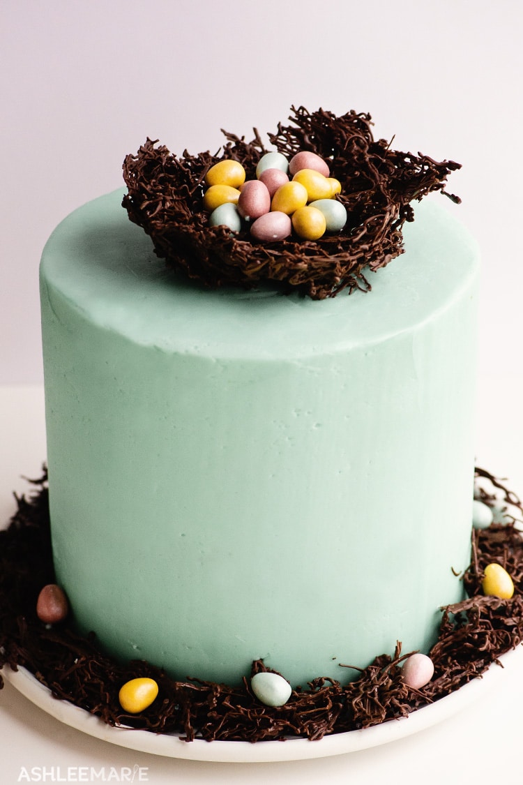A light blue frosted cake topped with a chocolate nest, full of chocolate candy eggs.