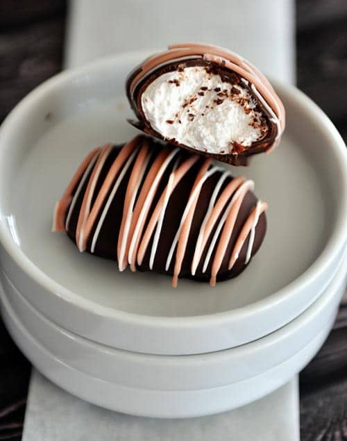 A chocolate covered egg on a plate drizzled with orange candy melts, and another leaning on top of it with a bite out of it showing marshmallow fluff in the middle.
