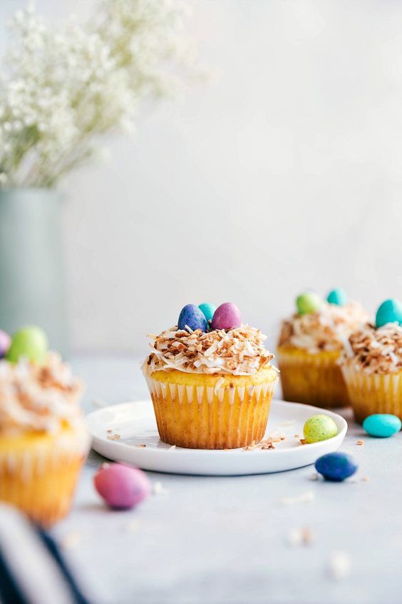 A yellow cupcake on a plate topped with white frosting, and toasted coconut to look like a birds nest, then three chocolate candies on top to look like eggs.