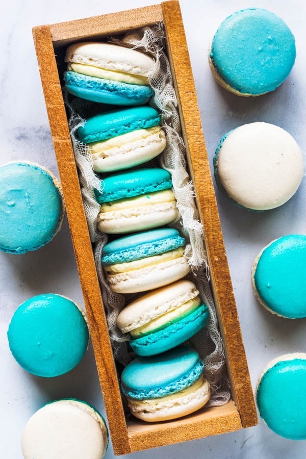 Macaroons in a box with a few on the sides. They have one half that is blue, and one half that is white and are filled with a white filling.