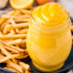 A jar full of a yellow, creamy mixture that is a homemade aioli, that is also on a plate with a bunch of french fries.