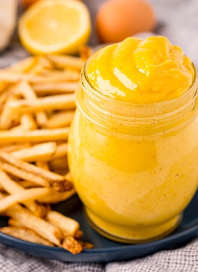 A jar full of a yellow, creamy mixture that is a homemade aioli, that is also on a plate with a bunch of french fries.