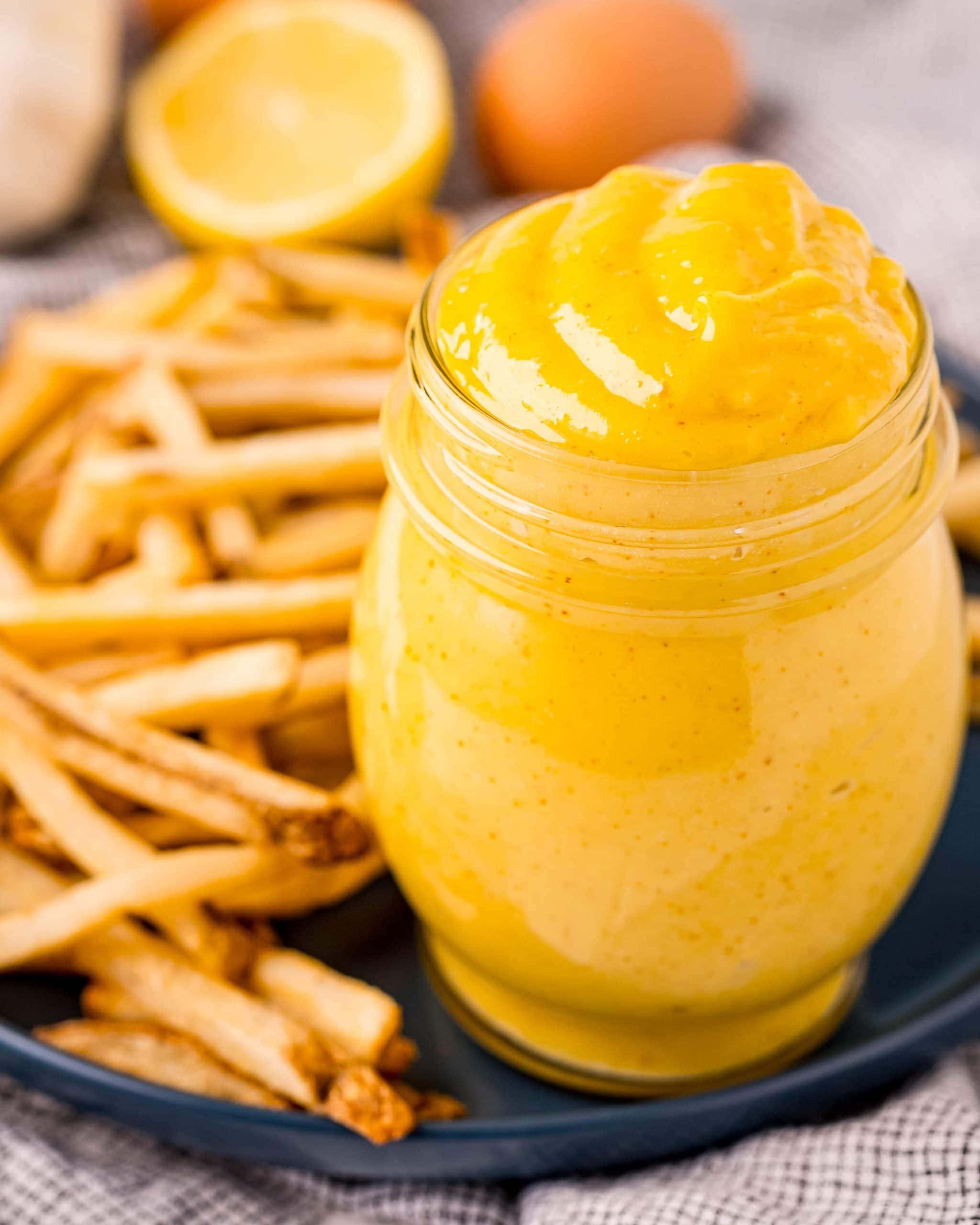 A jar full of a yellow, creamy mixture that is a homemade aioli, that is also on a plate with a bunch of french fries.