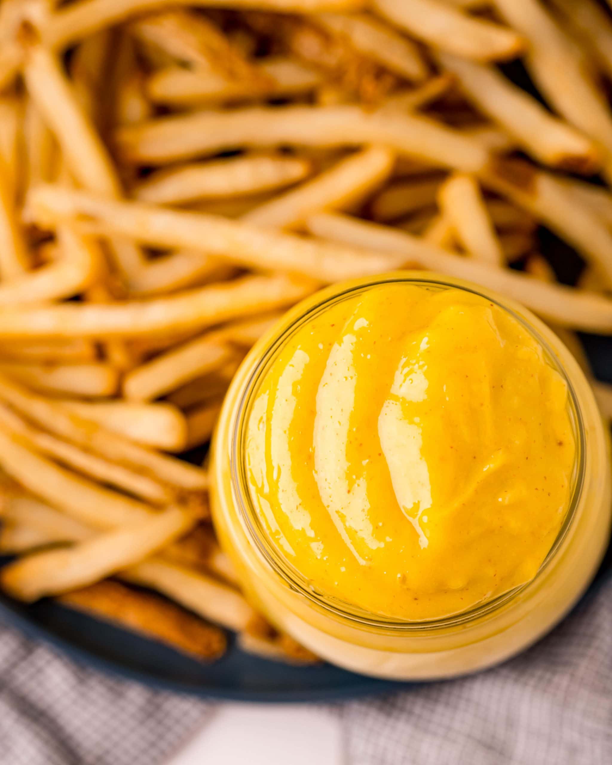 An overhead shot of a jar full of a yellow, creamy mixture that is a homemade aioli, that is also on a plate with a bunch of french fries.