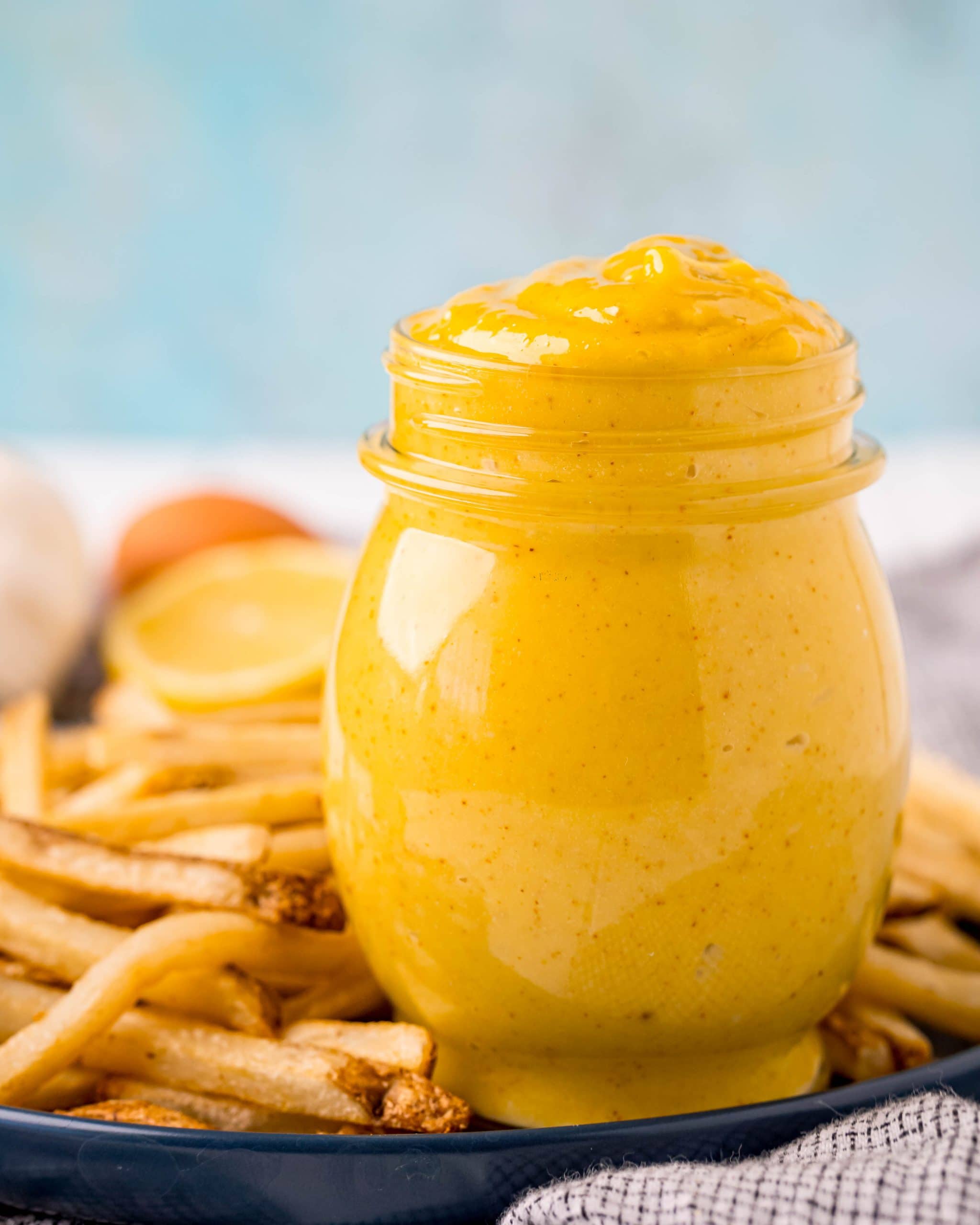 A jar full of a yellow, creamy mixture that is a homemade aioli, that is also on a plate with a bunch of french fries.