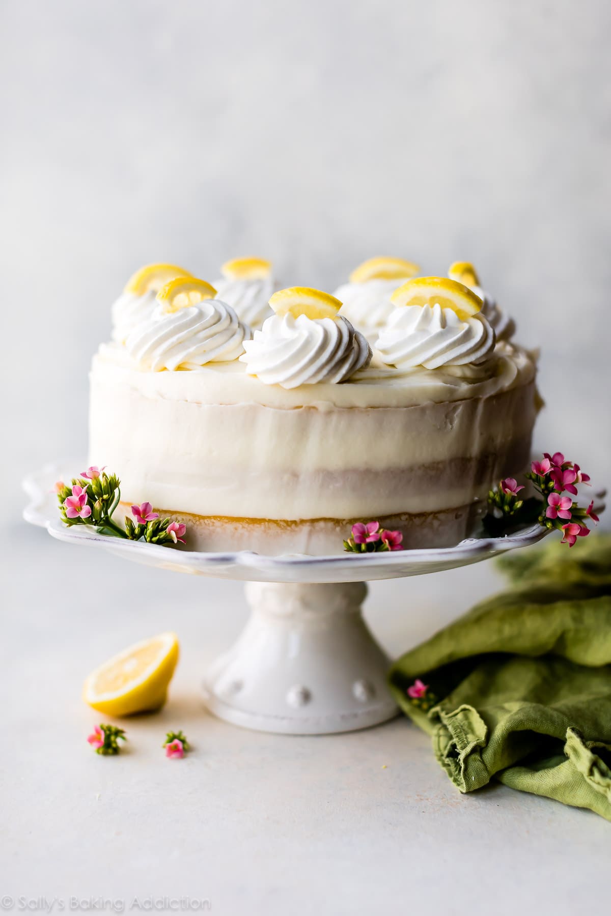 A layered lemon cake on a cake stand topped with a thin icing, and swirls of whipped cream and lemon slices.