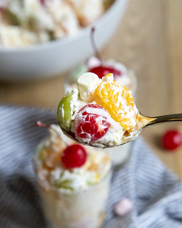 A spoonful of ambrosia salad with a maraschino cherry, green grape, marshmallow, and mandarin orange, above a bowl of the fruit salad.