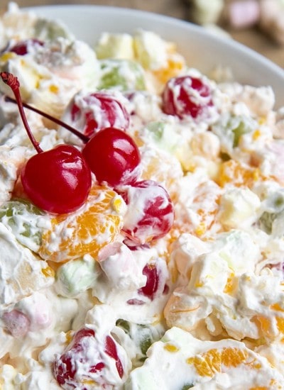 A close up of a bowl of fruit salad covered in a creamy sauce. Topped with two red maraschino cherries.