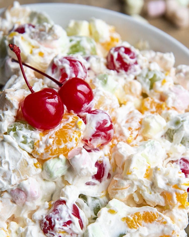 A close up of a bowl of fruit salad covered in a creamy sauce. Topped with two red maraschino cherries.
