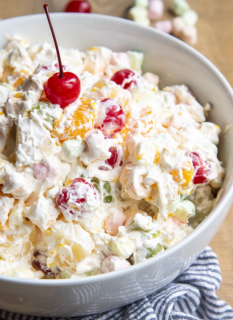 A close up of a bowl of fruit salad covered in a creamy sauce. Topped with a red maraschino cherries.