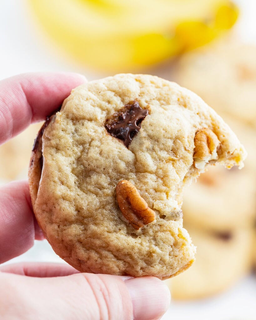 A hand holding a banana chocolate chip cookie with a bite out of it.