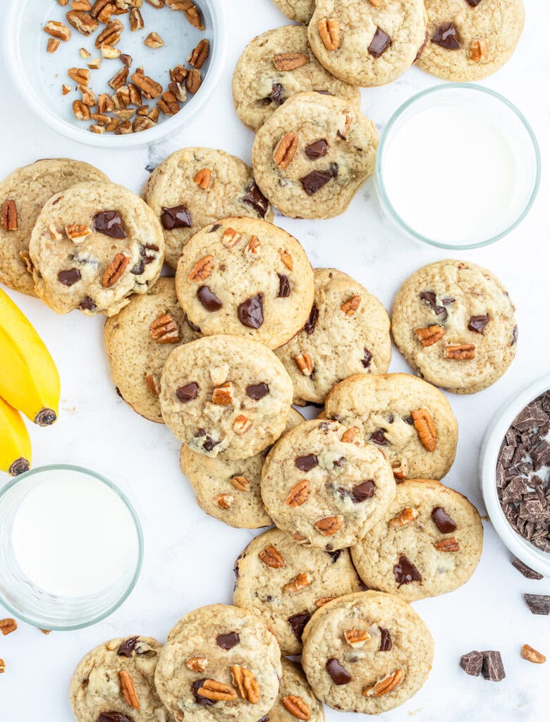 An above view of brown butter banana cookies full of walnuts, and chocolate chunks, next to glasses of milk.