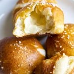 A plate with ball shaped soft pretzels, one is opened up, showing white melted cheese in the middle.