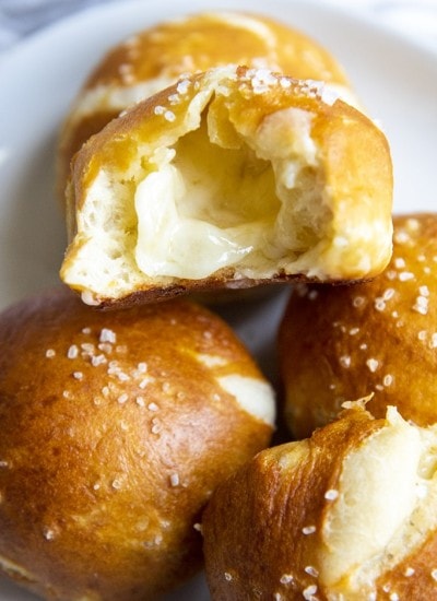 A plate with ball shaped soft pretzels, one is opened up, showing white melted cheese in the middle.