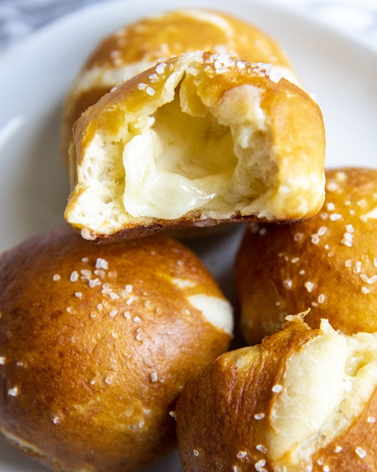 A plate with ball shaped soft pretzels, one is opened up, showing white melted cheese in the middle.