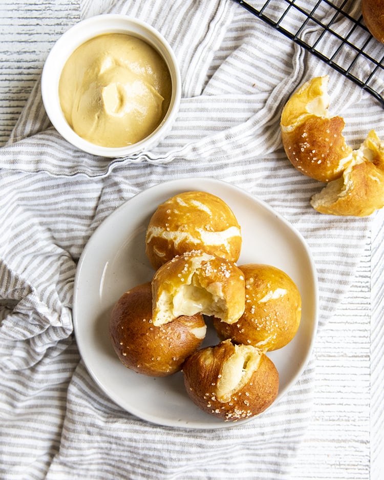 A small plate with cheese stuffed soft pretzels on it in a small pile. There is a bowl of mustard on the side, and another pretzel ball that is broken in half.