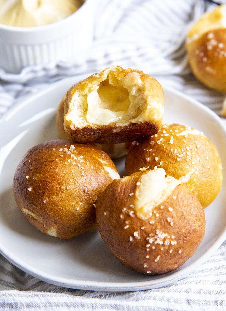 A plate with ball shaped soft pretzels, one is opened up, showing white melted cheese in the middle.