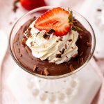 A glass jar full of chocolate pudding topped with whipped cream and half a strawberry.