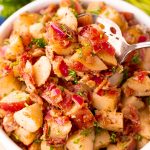 A close up of a bowl of warm german potato salad with red potatoes, bacon, and small pieces of red onion. There is a spoon in the bowl holding some of the salad.