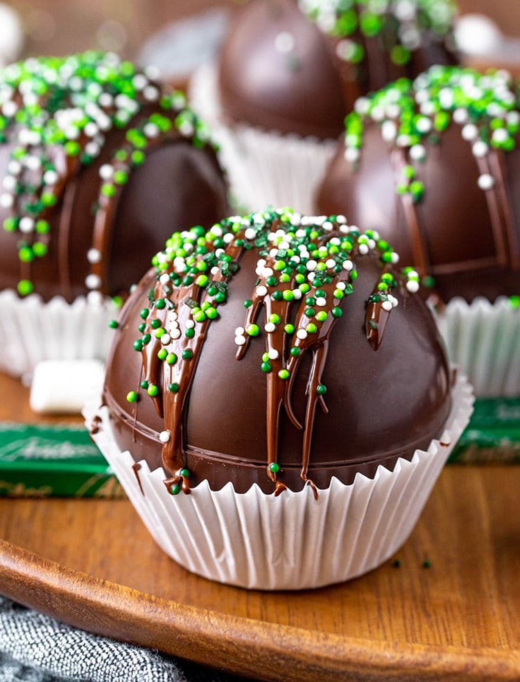 A ball shaped hot chocolate bomb topped with drizzled chocolate and green and white non pareil sprinkes. It it sitting in a muffin liner.