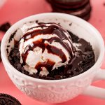 An Oreo mug cake in a white mug topped with a scoop of vanilla ice cream, drizzled with chocolate syrup.