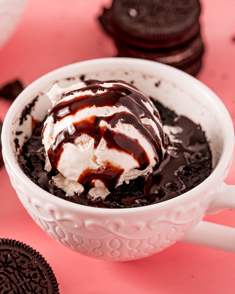 An Oreo mug cake in a white mug topped with a scoop of vanilla ice cream, drizzled with chocolate syrup.