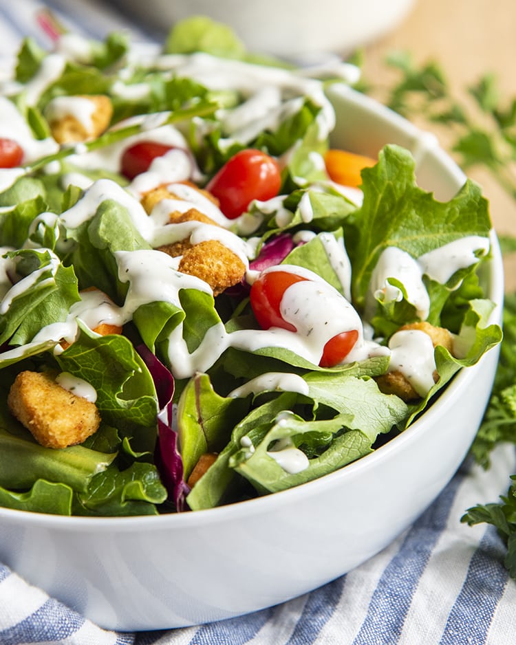 A white bowl full of salad topped with carrots, baby tomatoes, and croutons, and drizzled with ranch dressing.