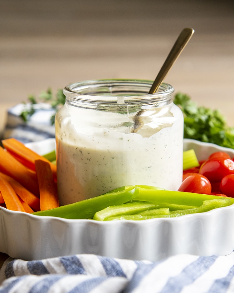 A clear jar full of homemade ranch dressing, about 3/4 with a spoon also in the jar. It is on a tray with sliced vegetables.