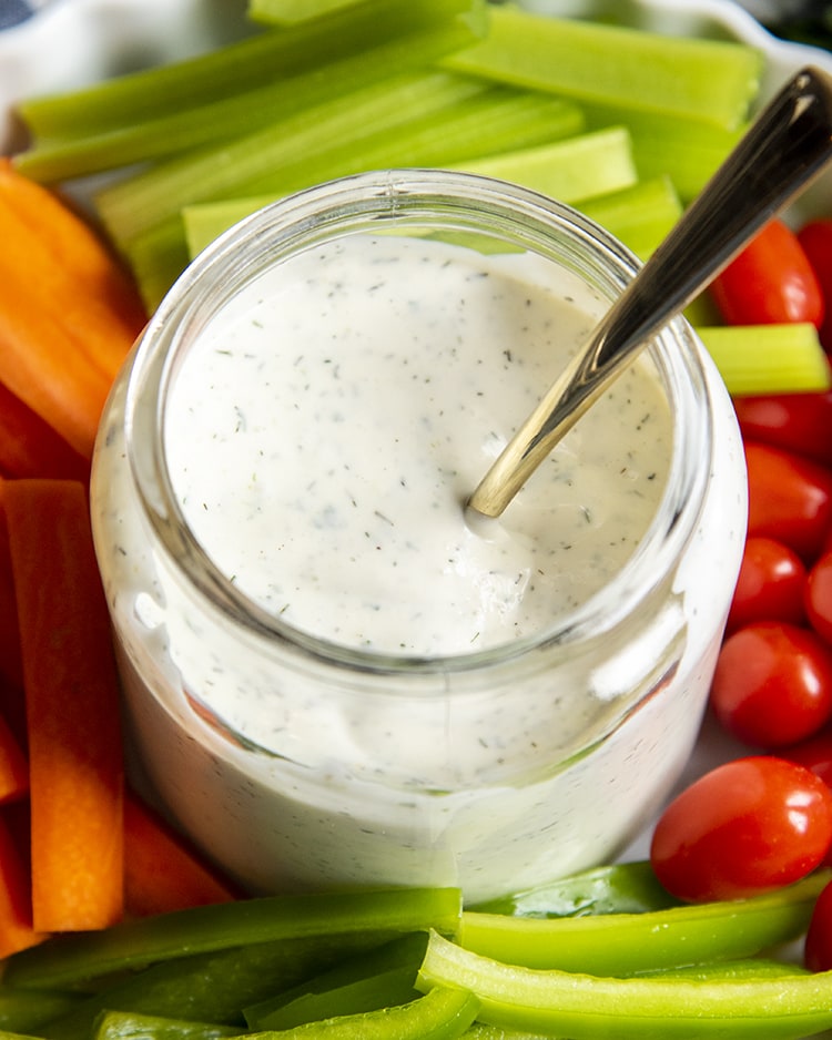 An overhead shot of a glass jar filled with ranch dressing with a golden spoon in the jar. The jar is surrounded by baby tomatoes, peppers, carrots, and celery.