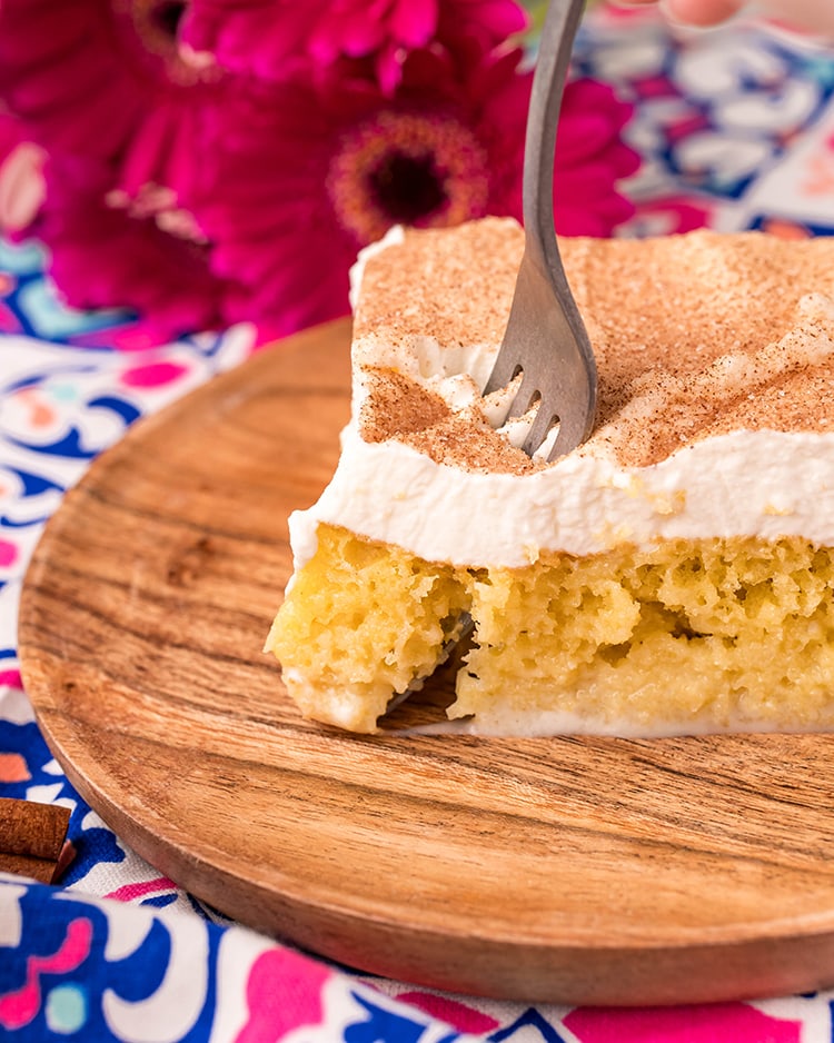 A piece of tres leches cake on a wooden plate, with a fork cutting a bite out of it.