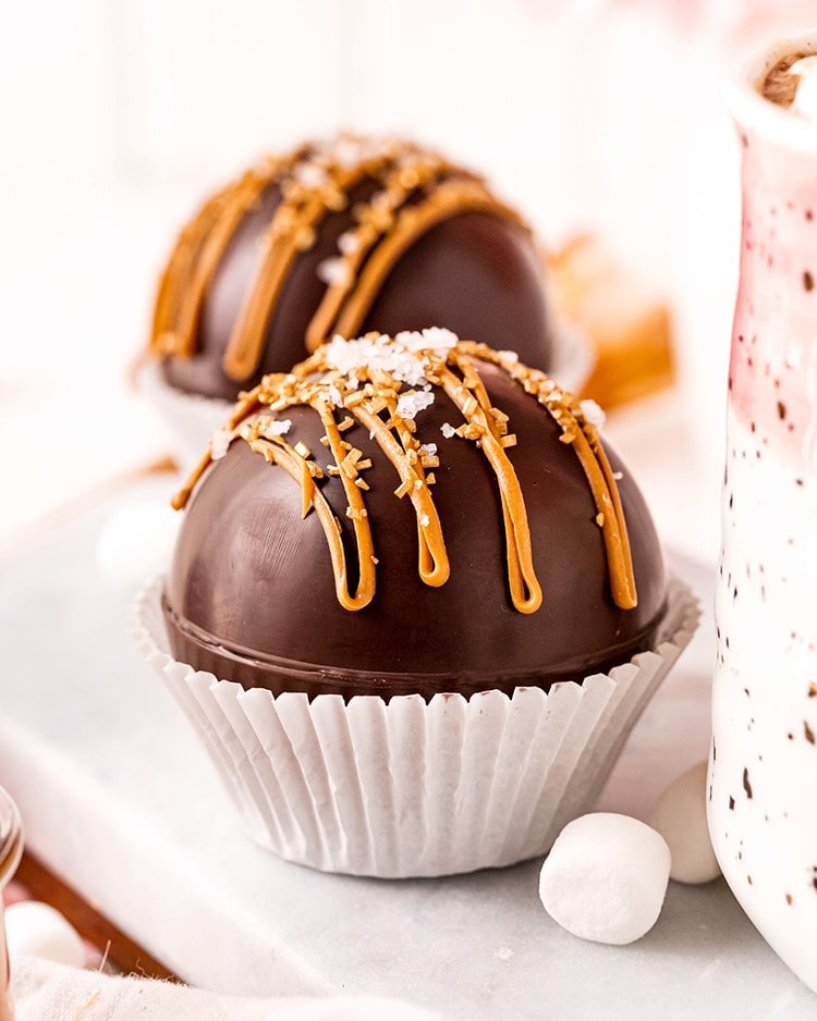 A hot chocolate bomb in a white paper muffin liner with caramel candy melts drizzled over the top, and sprinkled with sea salt.