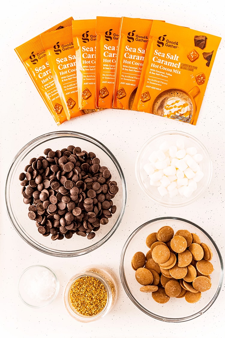 The ingredients needed to make caramel hot chocolate bombs. The packets of caramel hot chocolate, couverture chocolate in a bowl, mini marshmallows, caramel candy melts in a bowl, and two small bowls of golden sprinkles, and course flaky sea salt.