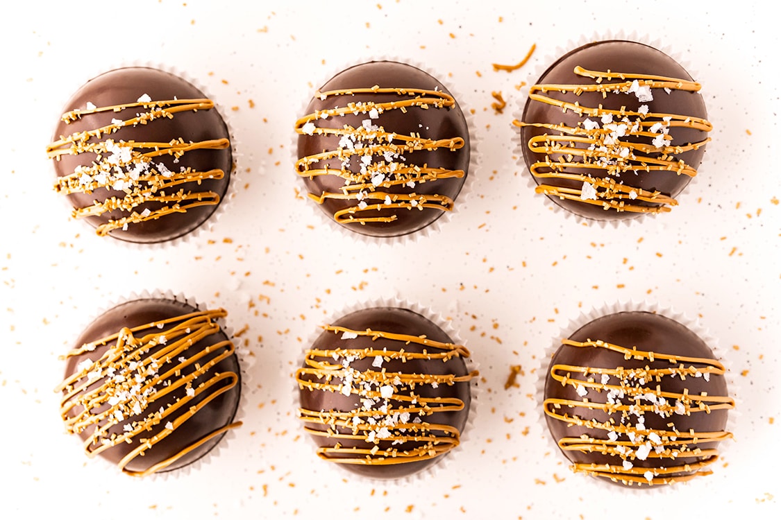 Six hot chocolate bombs arranged in two rows, they are chocolate domes with drizzles of a golden brown over the top, and sprinkled with flaky salt.