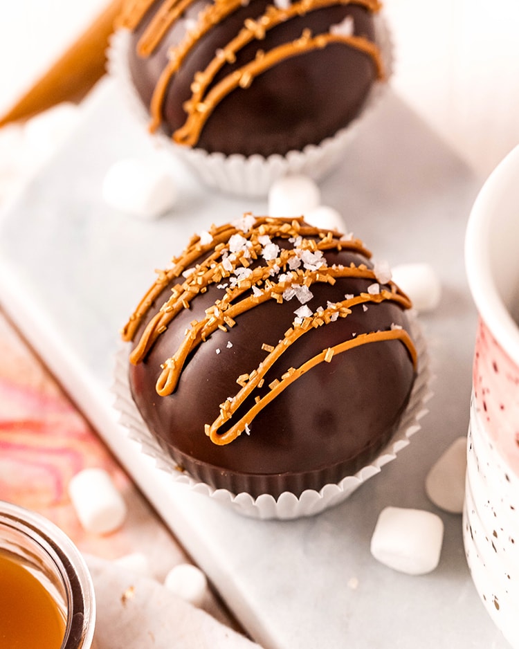 A hot chocolate bomb in a white paper muffin liner with caramel candy melts drizzled over the top, and sprinkled with sea salt.