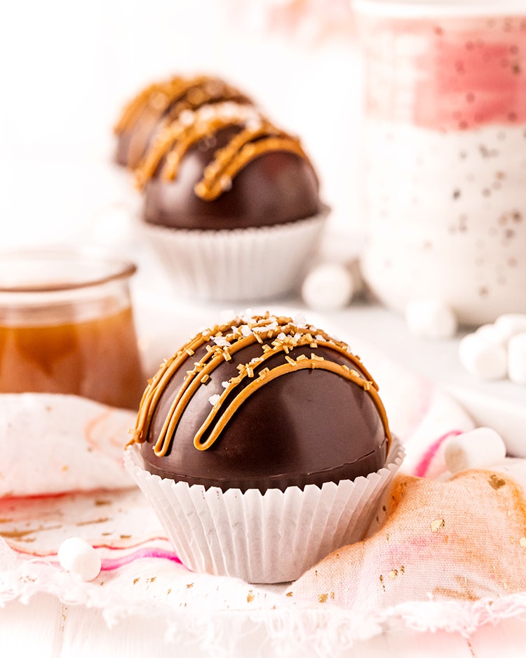 A hot chocolate bomb in a white paper muffin liner with caramel candy melts drizzled over the top, and sprinkled with sea salt. There are more hot chocolate mugs behind it too. 