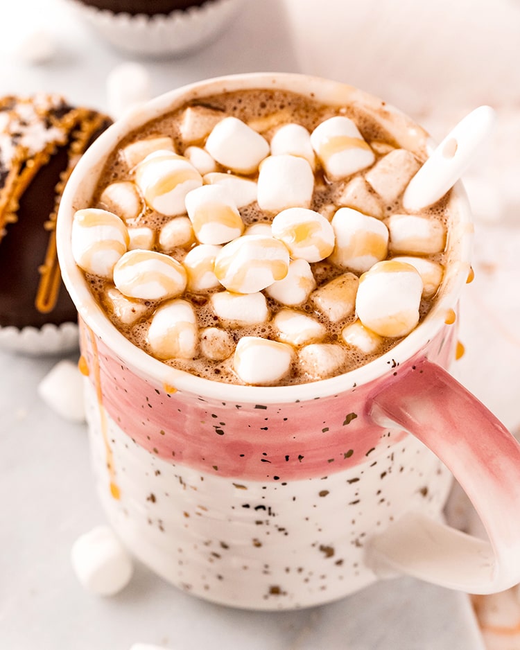A white and pink mug full of hot cocoa, and topped with mini marshmallows, and drizzled with caramel.