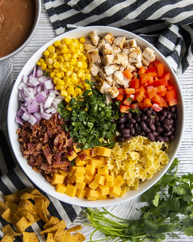 A bowl of the ingredients needed for bbq ranch pasta salad. There is farfalle noodles, black beans, red bell pepper, chopped chicken, corn, red onion, bacon, cheese cubes, and cilantro in the middle.