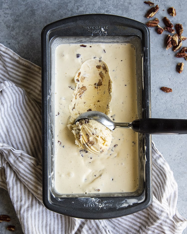A bread loaf pan full of a custard based butter pecan ice cream, with a scoop being pulled out of it. There are pecans showing through the ice cream.
