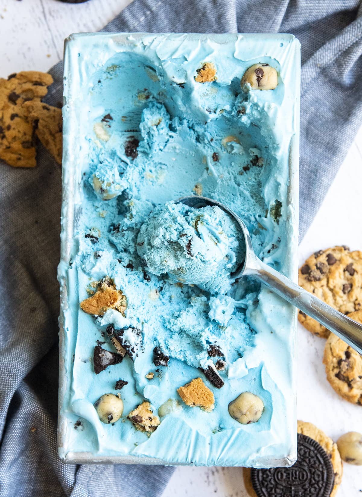 A container of blue cookie monster ice cream, that is full of chips ahoy cookies and oreo cookie pieces.