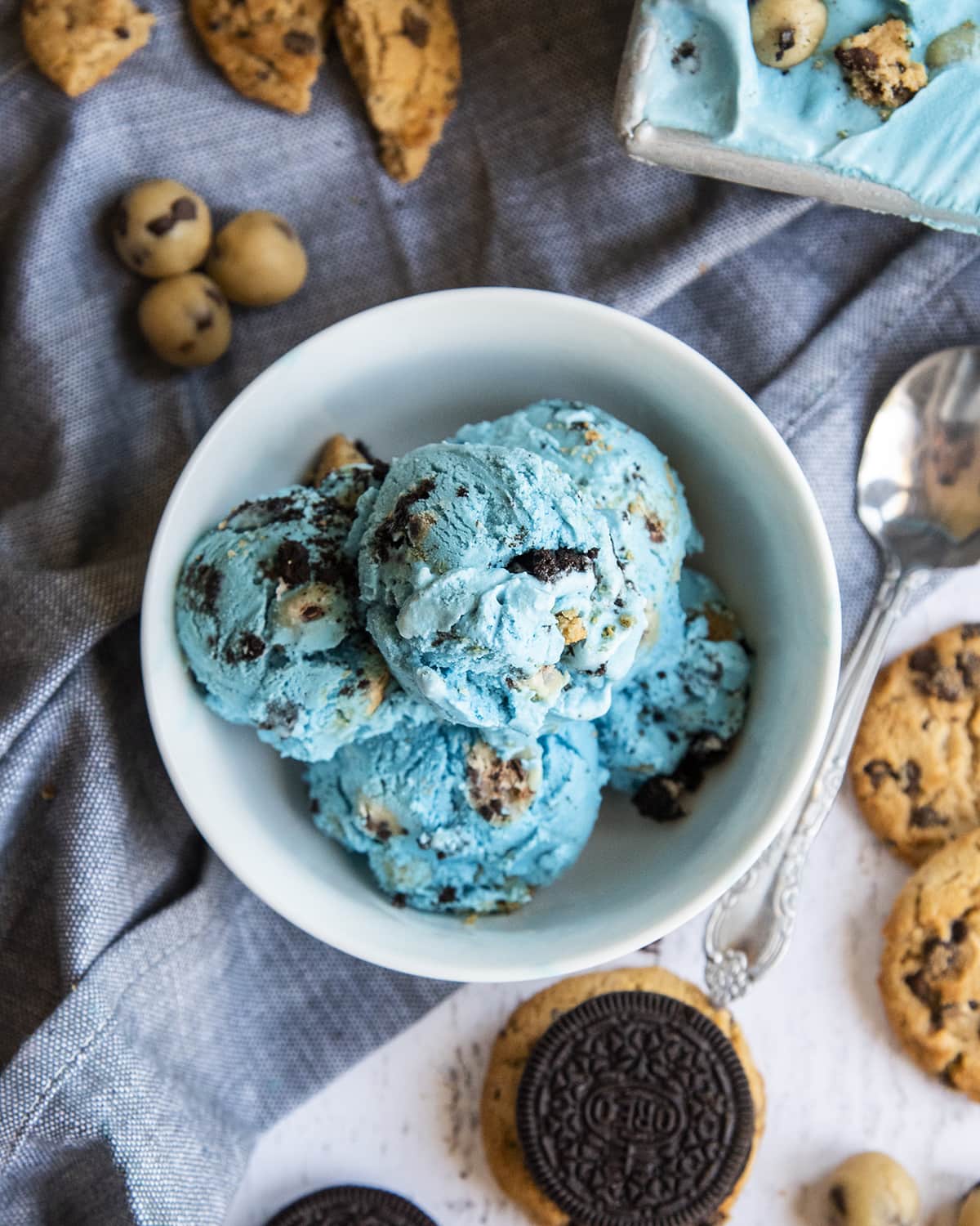 An overhead shot of a bowl of blue cookie monster ice cream full of Oreos, cookies, and cookie dough. Surrounded by chips ahoy cookies and Oreo cookies.