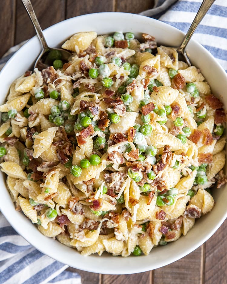 A close up of pasta salad with medium shells, bacon, and peas covered in a creamy white sauce.