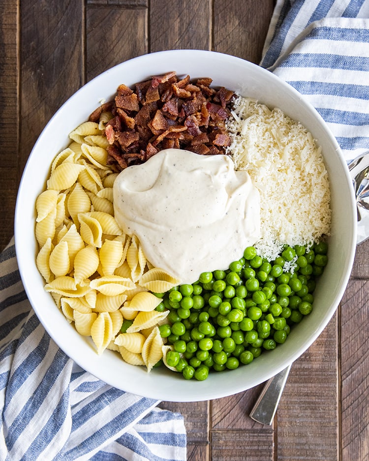 The ingredients needed to make creamy pasta salad, medium shells, crumbled bacon, grated parmesan, peas, and in the middle the white sauce.