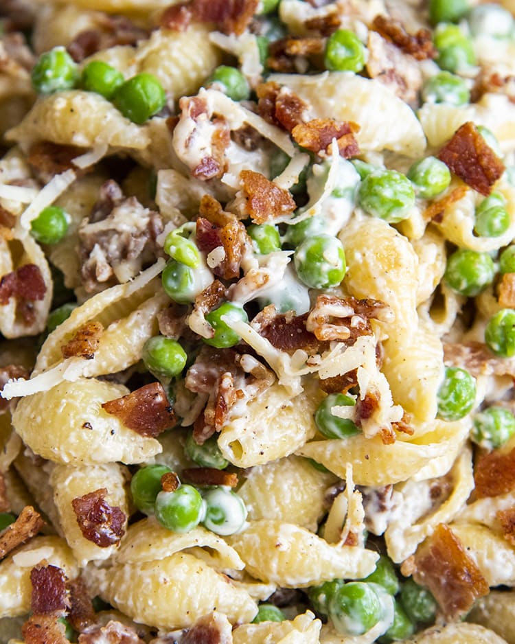 A close up of pasta salad with medium shells, bacon, and peas covered in a creamy white sauce.