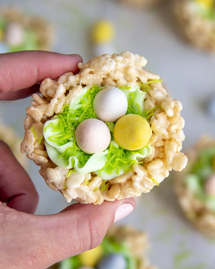 A hand holding an Easter Rice Krispie treat Nest, decorated to look like a nest with green frosting and chocolate eggs to look like a bird nest.