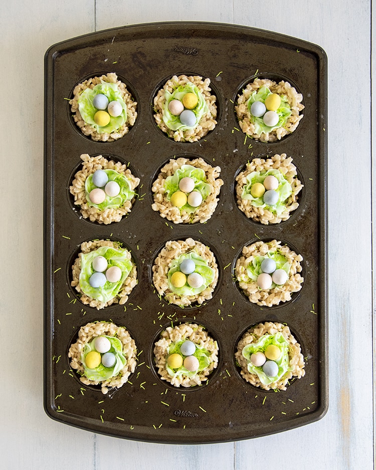 A muffin pan full of rice krispie treats decorated to look like nests with green frosting on top, and three chocolate eggs.