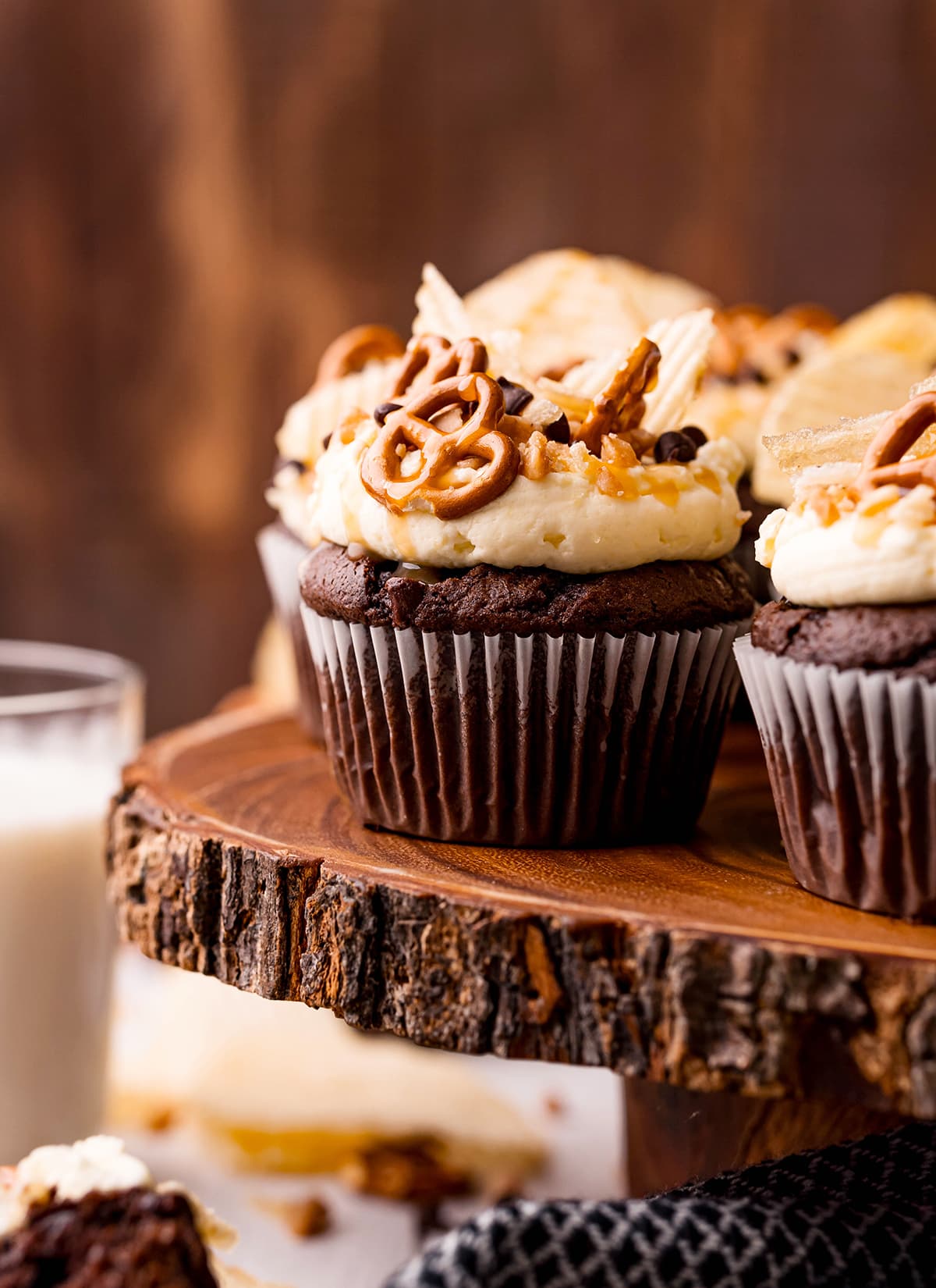 Chocolate cupcakes on a wooden stand topped with mini pretzels, ruffle chips, chocolate chips, and toffee pieces.