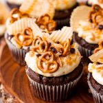 Chocolate cupcakes on a wooden stand topped with mini pretzels, ruffle chips, chocolate chips, and toffee pieces.