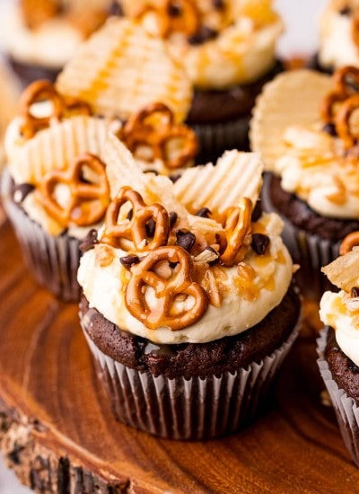 Chocolate cupcakes on a wooden stand topped with mini pretzels, ruffle chips, chocolate chips, and toffee pieces.